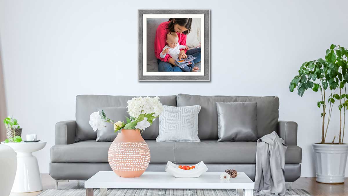 A simple photo of a mother reading a book with her baby, framed in a grey open grain wood frame above a grey settee